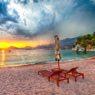 Budva yakınlarındaki Sveti Stefan 'da şezlonglar ve şemsiyelerle plajda dramatik bir gün batımı. Konum: Budva Riviera, Karadağ, Adriyatik denizi, Avrupa