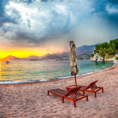 Budva yakınlarındaki Sveti Stefan 'da şezlonglar ve şemsiyelerle plajda dramatik bir gün batımı. Konum: Budva Riviera, Karadağ, Adriyatik denizi, Avrupa
