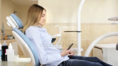 Genç bir kız bir dişçi sandalyede oturur ve kameraya bakarak bir smartphone kullanın