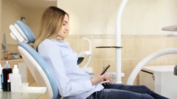 Uma jovem se senta em uma cadeira de dentista e usa um smartphone, olhando para a câmera — Vídeo de Stock