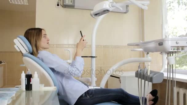 La chica sentada en la silla del dentista hace una selfie con un smartphone — Vídeo de stock