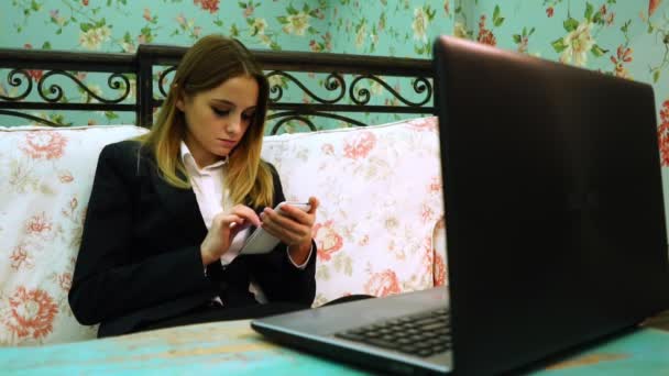 Joven mujer de negocios utiliza su teléfono en el sofá delante de la computadora portátil — Vídeos de Stock