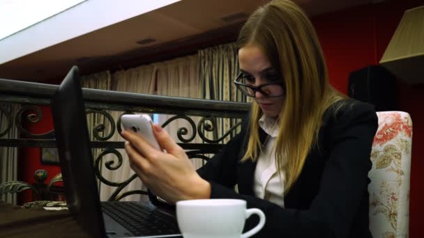 メガネと厳格なスーツ ビジネス女の子がノート パソコンの前でスマート フォンを使用します。 — ストック動画