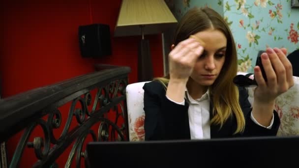 Молода дівчина в сорочці і куртці коригує свій макіяж перед онлайн записником зустрічей — стокове відео
