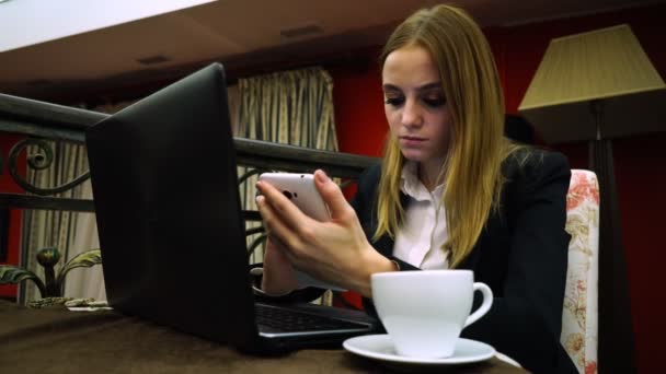 Uma menina em um café com um laptop e um smartphone sobre uma xícara de chá lê as notícias do telefone — Vídeo de Stock