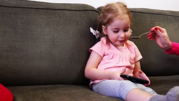 Девочка развлекается со своим смартфоном, пока ее кормят — стоковое видео