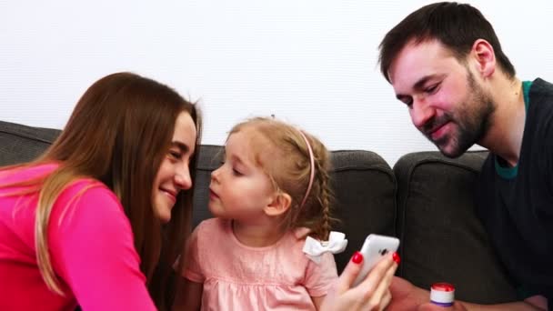 La familia mira el teléfono inteligente, la hija besa a mamá y papá — Vídeo de stock