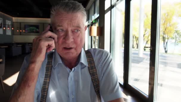 讲智能手机的老人慢吞吞地坐在窗边的咖啡店里. — 图库视频影像