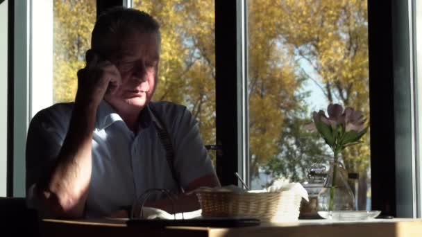 老人はスローモーションで窓の近くのカフェに座っているスマートフォンで話しています. — ストック動画