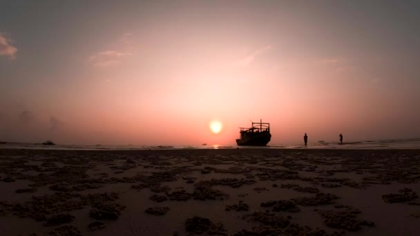 Солнце садится рядом с кораблем, пока люди прогуливаются по песчаному пляжу — стоковое видео