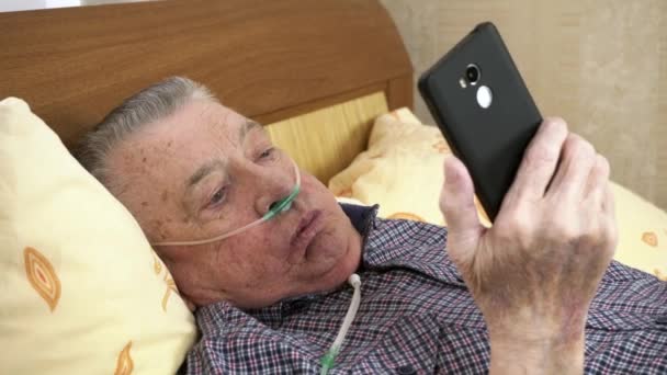 Viejo con cánula nasal está acostado en la cama sosteniendo el teléfono inteligente — Vídeos de Stock