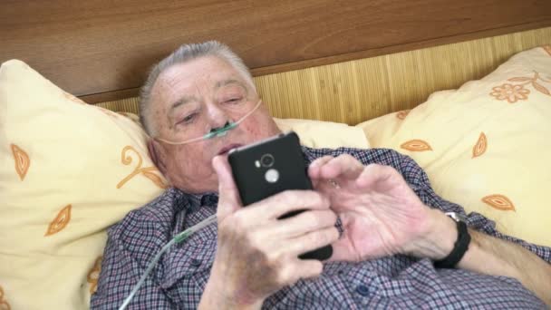 Viejo con cánula nasal está acostado en la cama con el teléfono en las manos — Vídeo de stock
