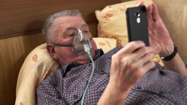 De man met het ademmasker ligt aan het bed en kijkt naar de telefoon. — Stockvideo