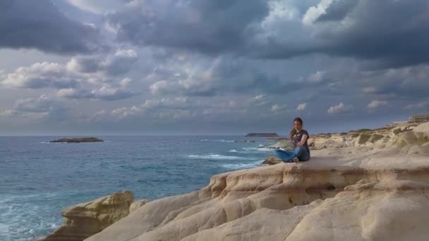 Una chica se sienta en una roca en la costa del mar — Vídeos de Stock