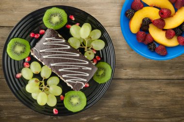 Kek ve meyve üzümleri, kivi, ahşap zemin üzerinde nar. Meyve manzaralı siyah tabakta tatlı.