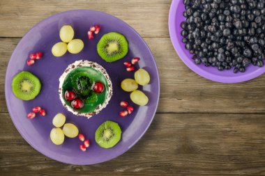 Kek ve meyve üzümleri, kivi, ahşap zemin üzerinde nar. Mor bir tabakta meyveli tatlı.