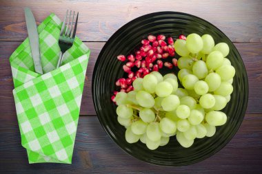 Üzüm, mor ahşap arka planda nar. Üzüm, nar, çatal ve bıçakla siyah bir tabakta.