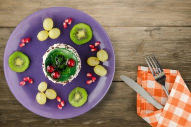 Kek ve meyve üzümleri, kivi, ahşap zemin üzerinde nar. Çatal ve bıçak manzaralı mor bir tabakta tatlı.