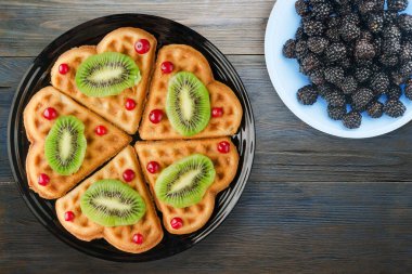 Mavi ahşap arka planda kivi ve kızılcıklı gofretler. Meyve manzaralı siyah tabakta waffle.