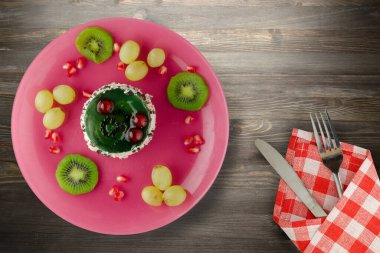 Kek ve meyve üzümleri, kivi, siyah ahşap zemin üzerinde nar. Çatal ve bıçak manzaralı pembe tepside meyveli tatlı.