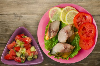 Sebze salatası manzaralı pembe tabakta tütsülenmiş uskumru. Kahverengi ahşap arka planda limonlu uskumru ve domates.