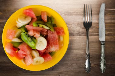 Kahverengi ahşap arka planda sağlıklı taze meyve salatası tabağı. Çatallı ve bıçak manzaralı meyve salatası.