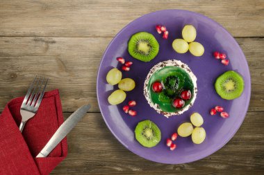 Kek ve meyve üzümleri, kivi, ahşap zemin üzerinde nar. Çatal ve bıçak manzaralı mor bir tabakta tatlı.