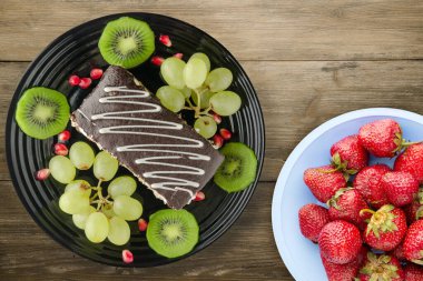 Kek ve meyve üzümleri, kivi, ahşap zemin üzerinde nar. Meyve manzaralı siyah tabakta tatlı.