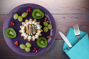 Kek ve meyve üzümleri, kivi, mor ahşap arka planda nar. Çatal ve bıçak manzaralı mor bir tabakta tatlı.