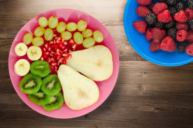 Meyve karışımı armut, kivi, üzüm, muz, kahverengi ahşap zemin üzerinde nar. Pembe bir tabakta meyveler.