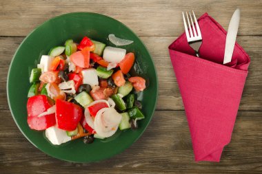 Kahverengi ahşap arka planda Yunan salatası. Çatal ve bıçak manzaralı yeşil tabakta Yunan salatası. sağlıklı yiyecekler