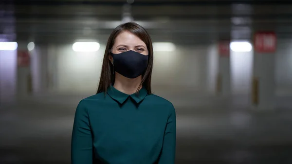 Happy woman smiles in a black mask in an underground parking lot, protecting against virus and infections during an epidemic.