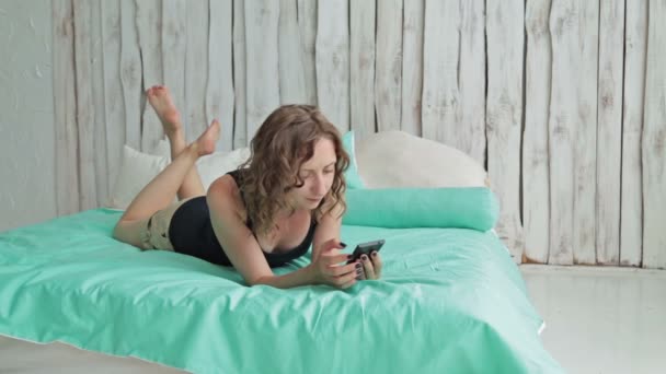 Young curly woman lying in the bed with smartphone — Stock Video