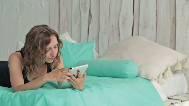 Mujer rizada joven acostada en la cama con la tableta pc. Dolly disparó — Vídeos de Stock