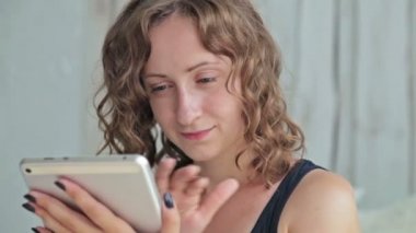 Genç kıvırcık kadın yatakta tablet pc ile oturuyordu. Dolly vurdu