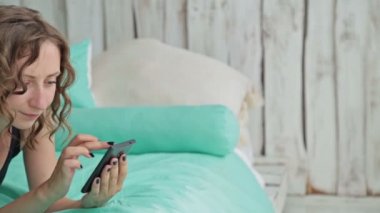 Smartphone ile yatakta yatan genç kadın kıvırcık. Dolly vurdu
