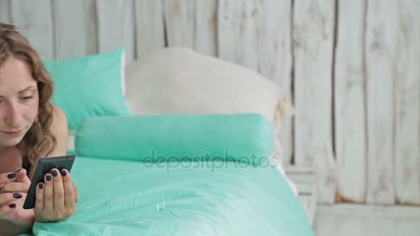 Jeune femme bouclée couchée dans le lit avec smartphone. Dolly shot — Video