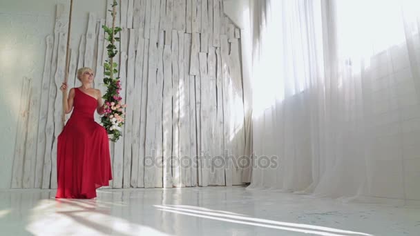 Όμορφη, νεαρή και αισθησιακή γυναίκα σε κόκκινο φόρεμα σε μια κούνια διακοσμημένο με λουλούδια — Αρχείο Βίντεο