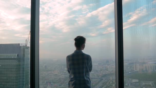 Mujer pensativa mirando paisaje urbano a través de la ventana del rascacielos - vista trasera — Vídeo de stock