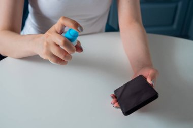 Dezenfeksiyon, koruma, önleme, COVID 19, koronavirüs, güvenlik, sağlık ve teknoloji konsepti. Kadın antiseptik püskürtüyor, beyaz masaya siyah akıllı telefon temizliyor.