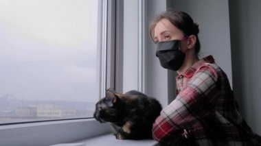 Tıbbi maskeli kadın ve pencereden dışarı bakan siyah kedi. Ağır çekim.