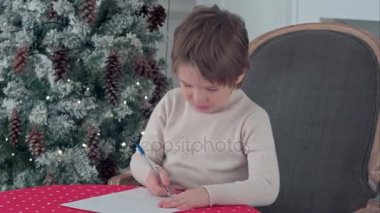 Küçük çocuk masada Noel Baba'ya mektup yazmaya konsantre