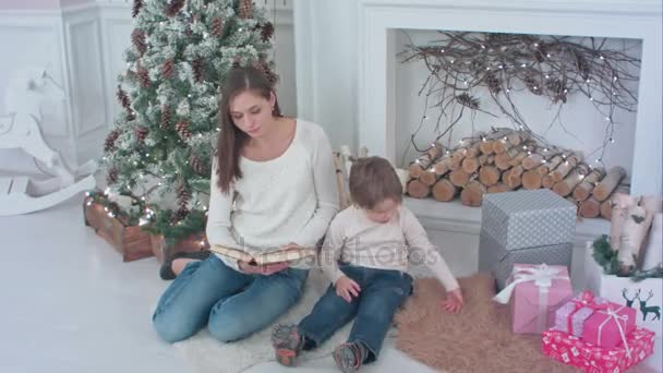 Молода мама читання книги, щоб відволікти її сина від Різдва представляє — стокове відео