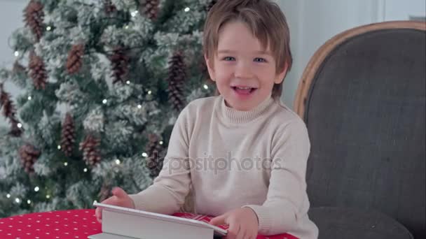 Uśmiechnięty chłopiec dziecko siedzi na krześle i grając z tabletu w czasie Bożego Narodzenia — Wideo stockowe