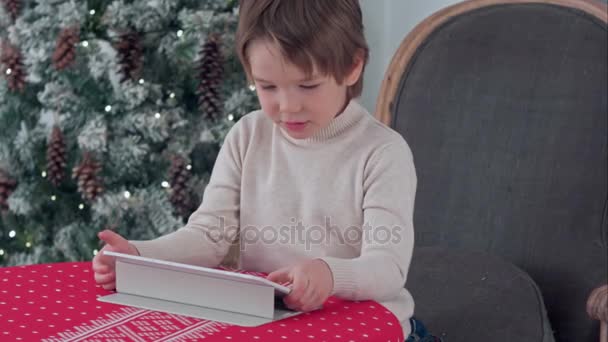 Lindo niño usando tableta mientras está sentado en un sillón grande en casa sobre el fondo del árbol de Chirstmas — Vídeos de Stock