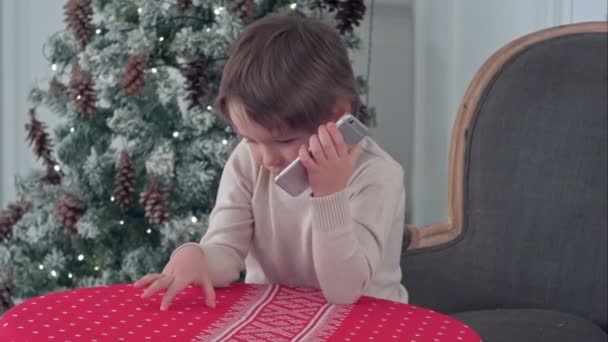Schattige kleine jongen roepen Santa zittend op een grote fauteuil thuis over Chirstmas boom achtergrond — Stockvideo