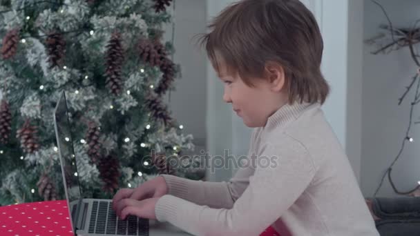 Lächelnder kleiner Junge tippt Brief an Weihnachtsmann auf Laptop neben dem Weihnachtsbaum — Stockvideo