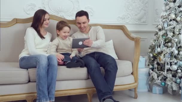 Pappa, Mamma och deras lilla son ha roligt genom att spela tillsammans med en tablett som sitter på en soffa — Stockvideo