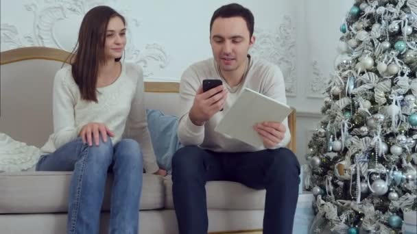 Jong koppel chillen op de Bank met de man bedrijf telefoon en tablet en praat met de camera — Stockvideo