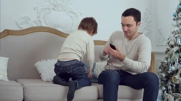 Pequeño hijo saltando en el sofá para distraer a su ocupado padre hablando por teléfono — Vídeo de stock